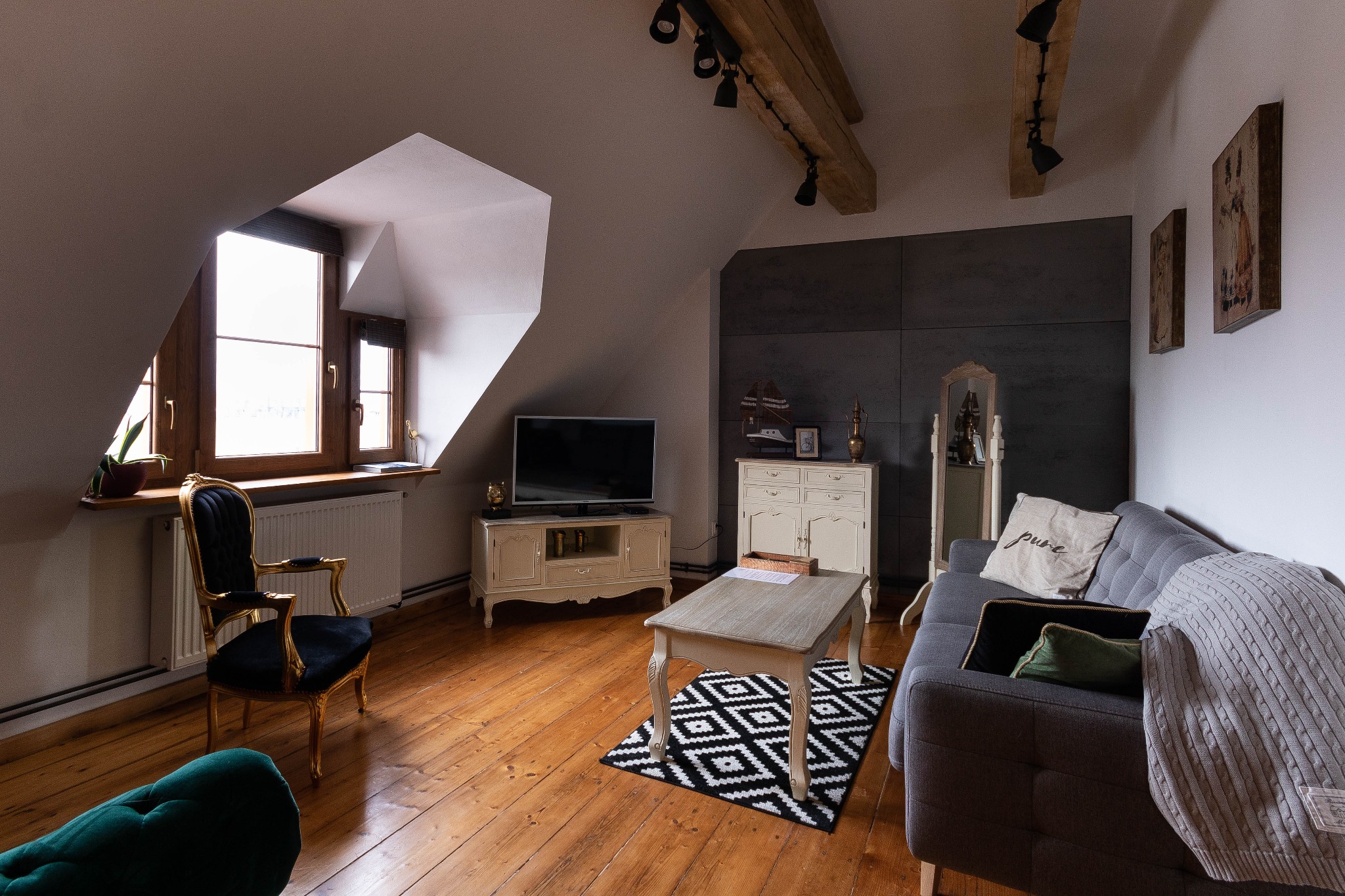Salon na poddaszu z drewnianą podłogą, szarą sofą, czarnym fotelem z pozłacanymi zdobieniami, geometrycznym dywanem i drewnianymi belkami stropowymi z oświetleniem punktowym.