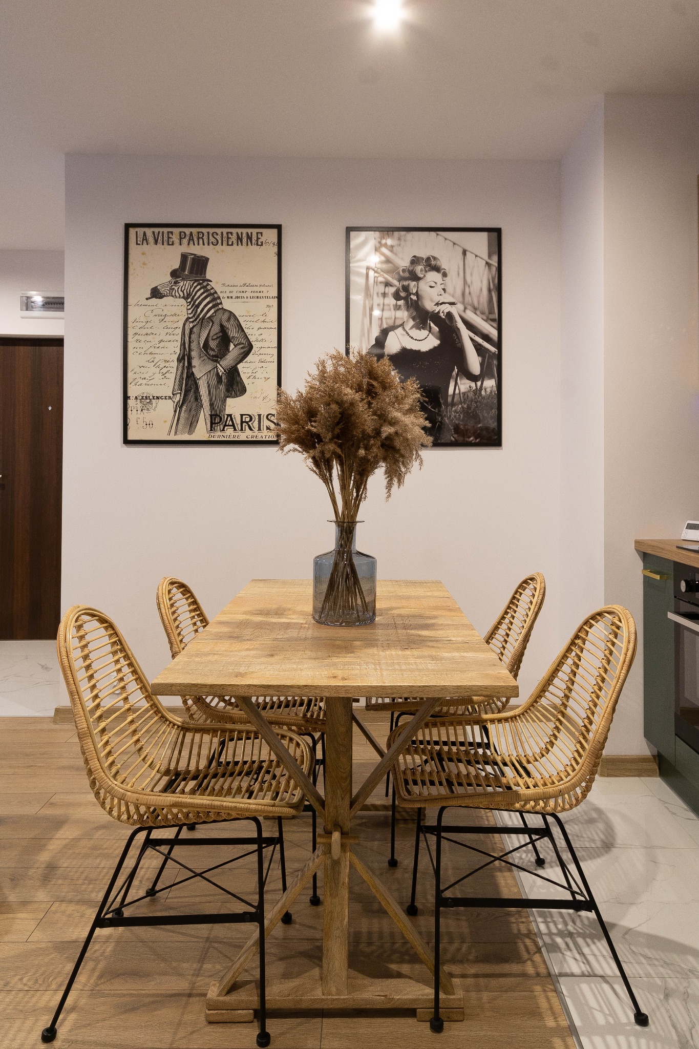 Jadalnia z drewnianym stołem, rattanowymi krzesłami i wazonem z suszonymi trawami. Na ścianie wiszą plakaty w czarno-białych ramach.