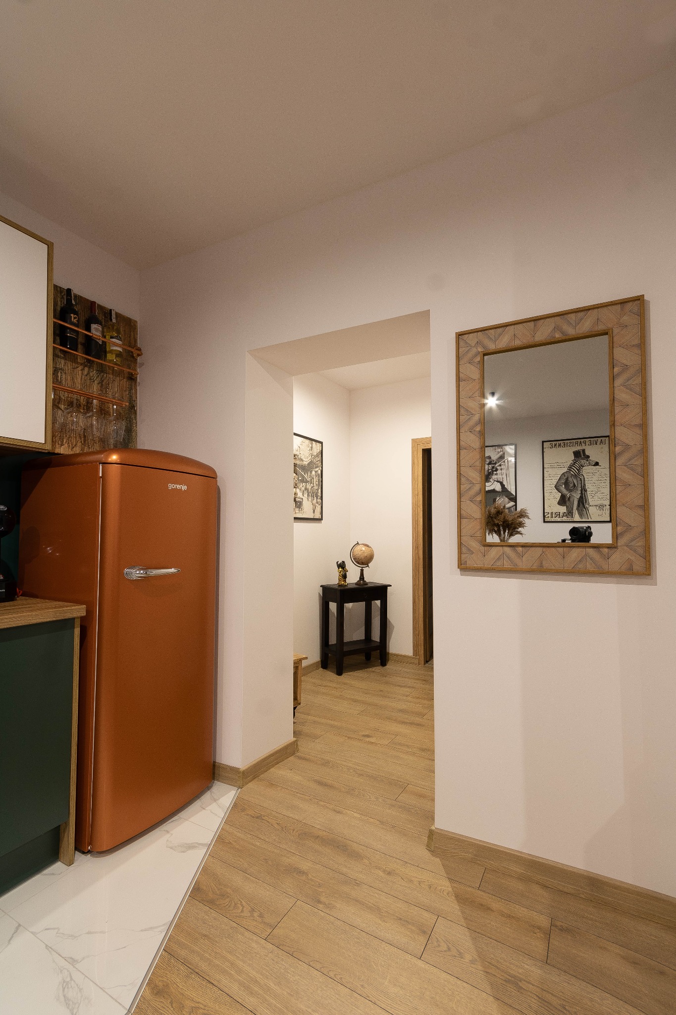 Wnętrze mieszkania z pomarańczową lodówką retro, drewnianą podłogą i lustrem z geometryczną ramą odbijającym plakat w korytarzu.