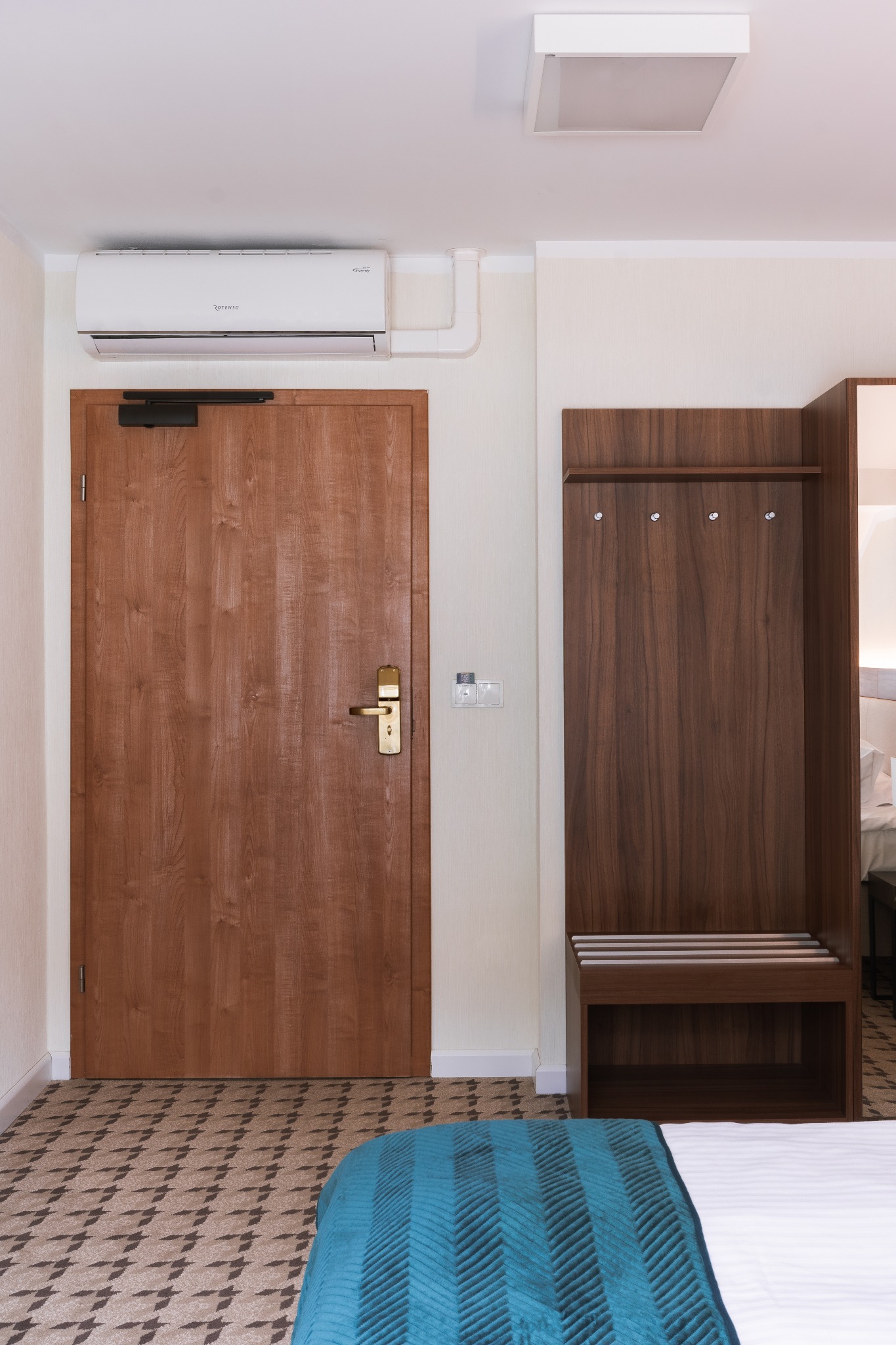 Wnętrze pokoju hotelowego z drewnianymi drzwiami, wieszakiem na ubrania z siedziskiem i klimatyzacją na ścianie.