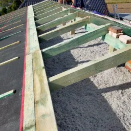 Wypełnienie przestrzeni między krokwiami wełną mineralną, widoczna konstrukcja drewniana dachu z zielonymi belkami, czarna membrana dachowa z czerwoną taśmą uszczelniającą, fragment dachu...