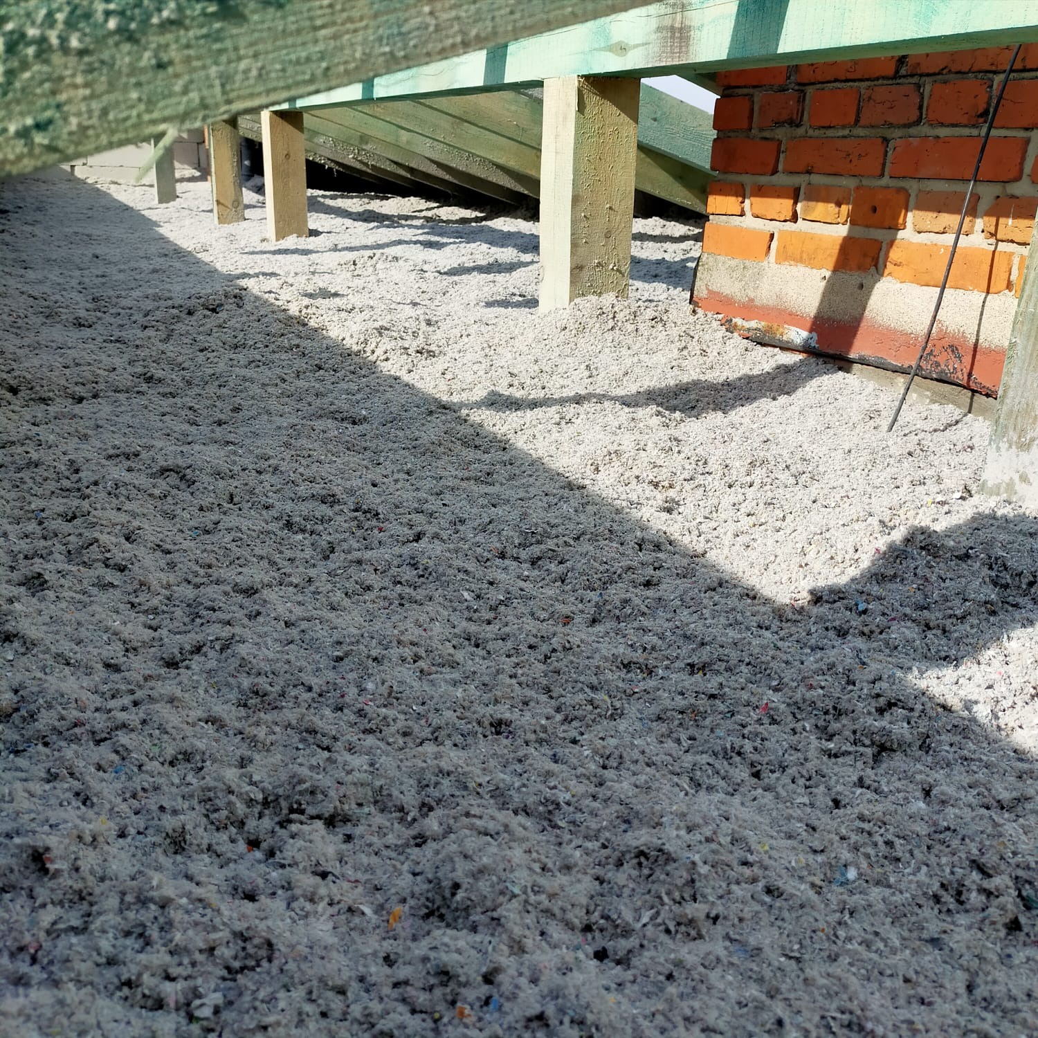 Poddasze z drewnianą konstrukcją i ceglanym kominem wypełnione szarą, włóknistą izolacją w trakcie prac ociepleniowych, widoczne drobne kolorowe elementy w izolacji.