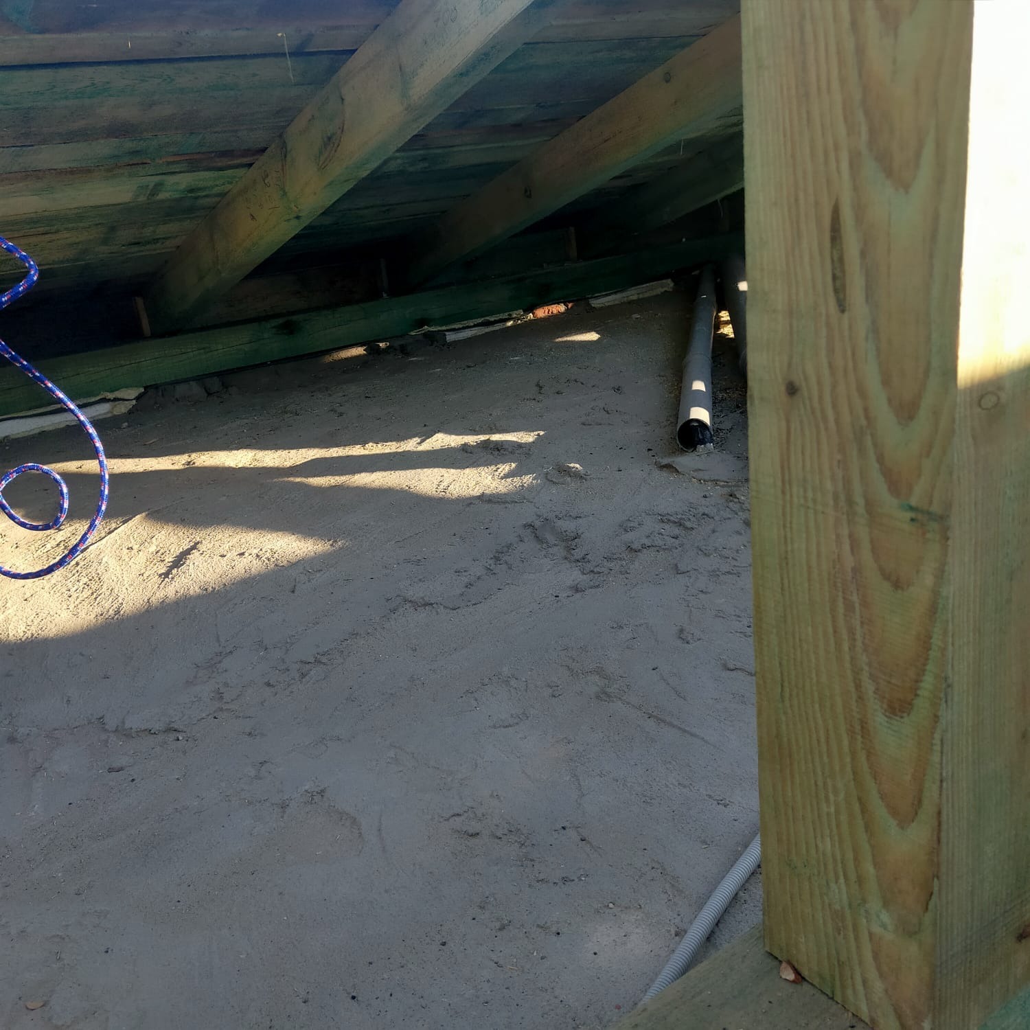 Poddasze w trakcie ocieplania: widoczne drewniane krokwie, szara izolacja częściowo ułożona, rury instalacyjne i fragment drewnianego słupa konstrukcyjnego.