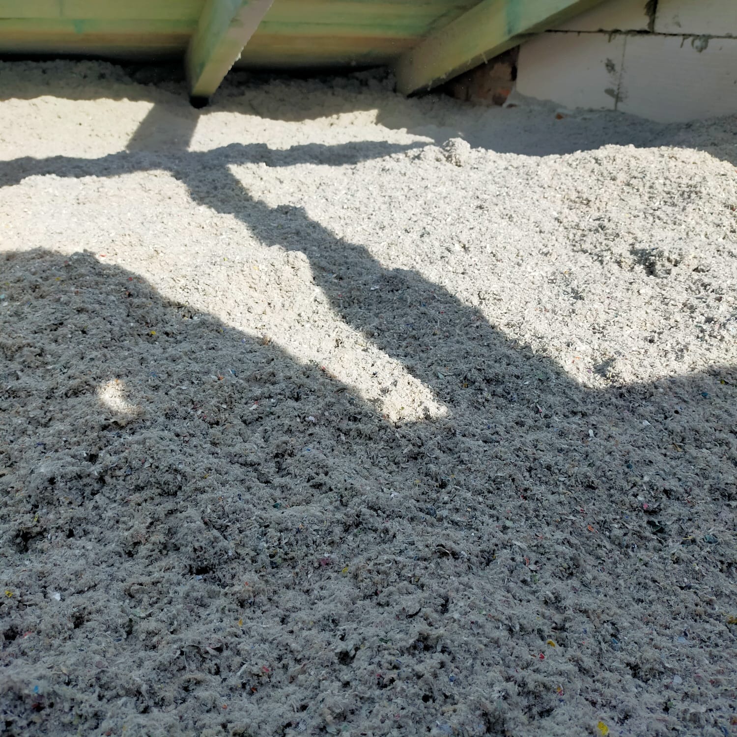 Szara, sypka izolacja z widocznymi kolorowymi elementami, ułożona pod drewnianą konstrukcją dachu i ścianą z bloczków betonowych, oświetlona promieniami słońca tworzącymi cienie.