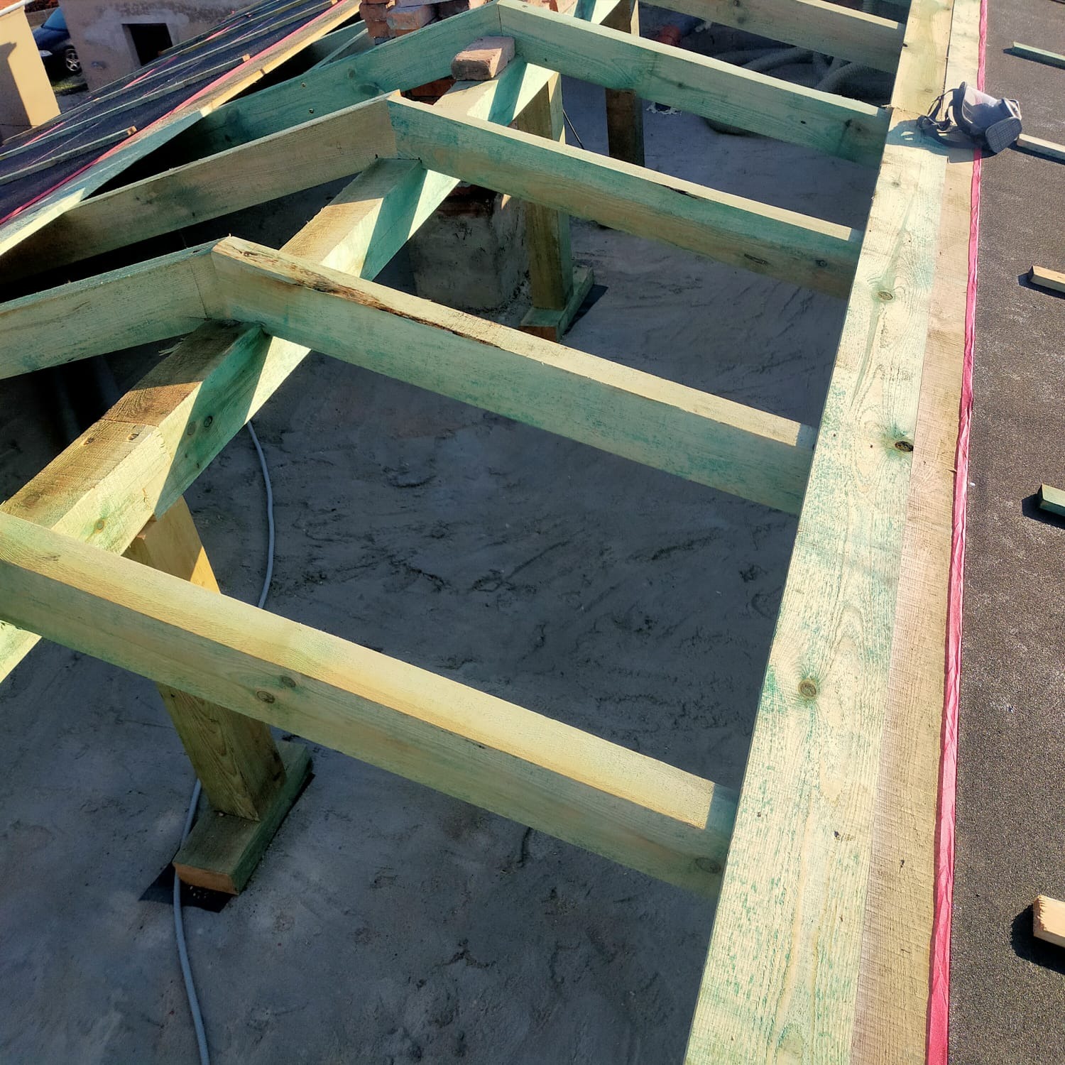 Konstrukcja drewniana dachu poddasza z impregnacją na zielono, widoczne elementy więźby dachowej na podłożu z papy i resztek materiałów budowlanych.