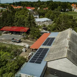 Widok z góry na instalację fotowoltaiczną na dachu budynku gospodarczego z szarej cegły, obok budynek z czerwonym dachem, w tle zabudowa wiejska i zieleń.
