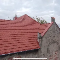 Czerwona dachówka ceramiczna na budynku z elementami architektonicznymi. Widoczny komin i fragmenty elewacji z dekoracyjnymi detalami. Tło stanowi pochmurne niebo.