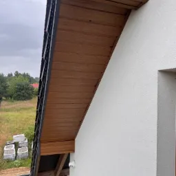 Drewniana podbitka dachowa z czarnymi elementami wykończeniowymi, widoczna z boku budynku z białą elewacją. W tle zieleń drzew i trawy, oraz ułożone kostki brukowe.