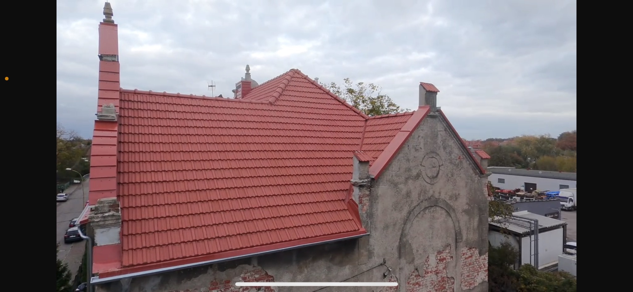 Czerwona dachówka ceramiczna na budynku z elementami architektonicznymi. Widoczny komin i fragmenty elewacji z dekoracyjnymi detalami. Tło stanowi pochmurne niebo.