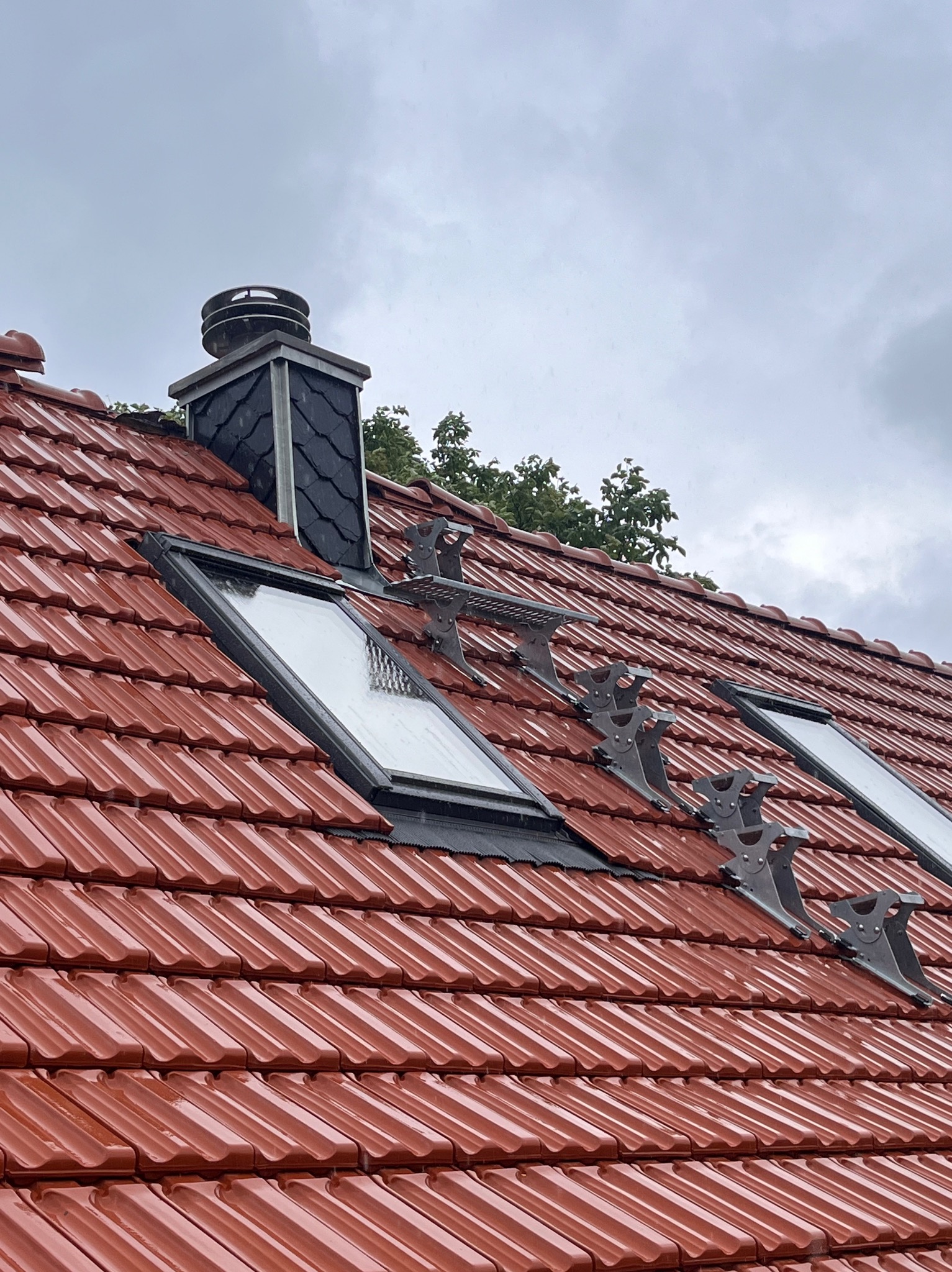 Czerwona dachówka ceramiczna z oknami dachowymi i zabezpieczeniami przeciwśniegowymi. Widoczny komin z czarną obudową. Pochmurne niebo w tle.