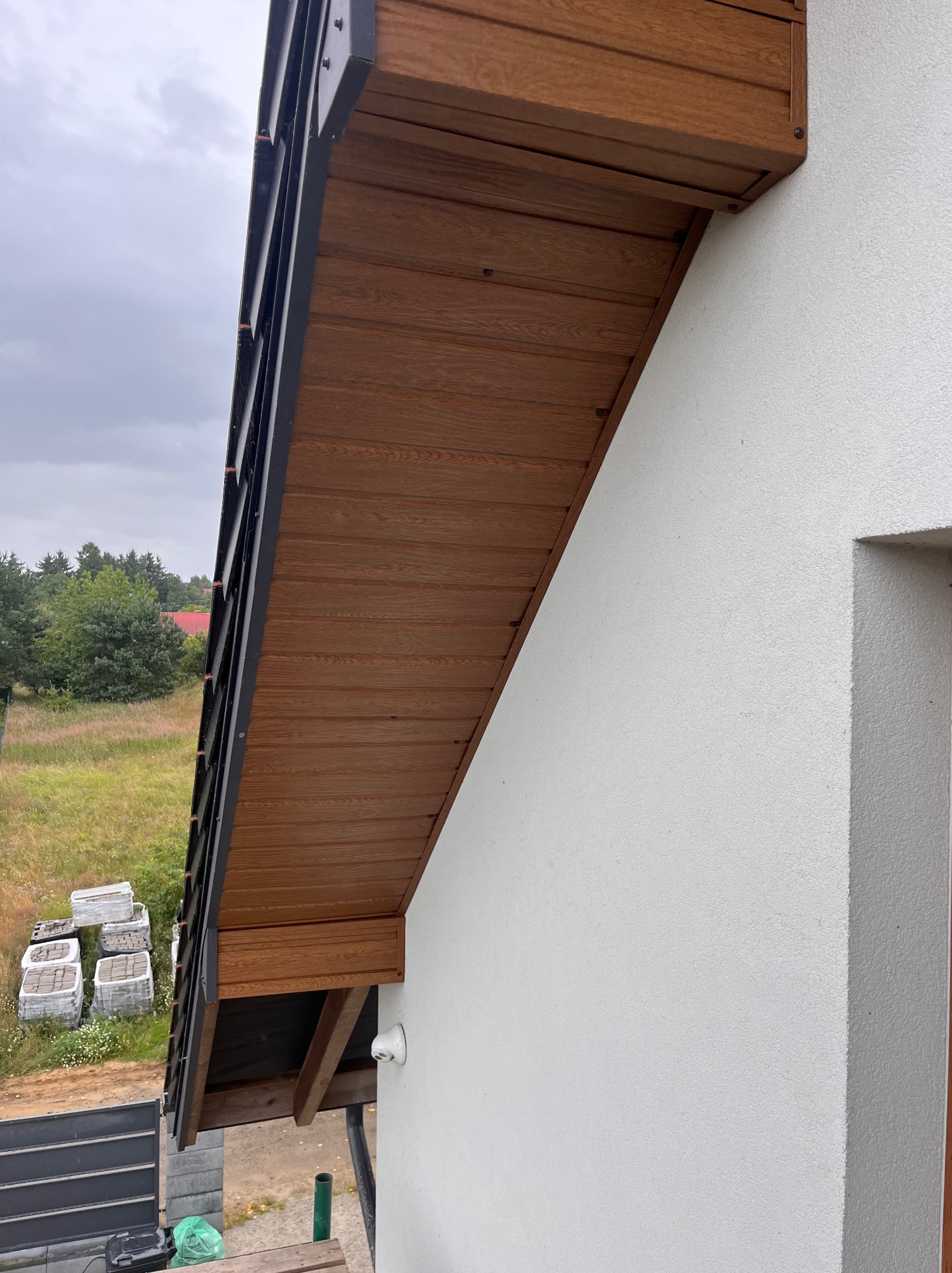 Drewniana podbitka dachowa z czarnymi elementami wykończeniowymi, widoczna z boku budynku z białą elewacją. W tle zieleń drzew i trawy, oraz ułożone kostki brukowe.