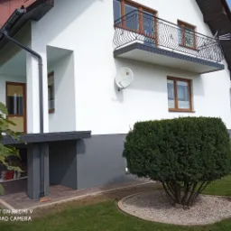 Dwupiętrowy dom z białymi ścianami, brązowymi oknami i balkonem z czarną balustradą, widok z zewnątrz na elewację i starannie przycięty krzew przed domem.