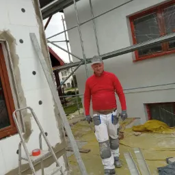 Pracownik w czerwonej bluzie na tle rusztowania i ściany budynku z zamontowanym styropianem, przygotowujący się do dalszych prac elewacyjnych.