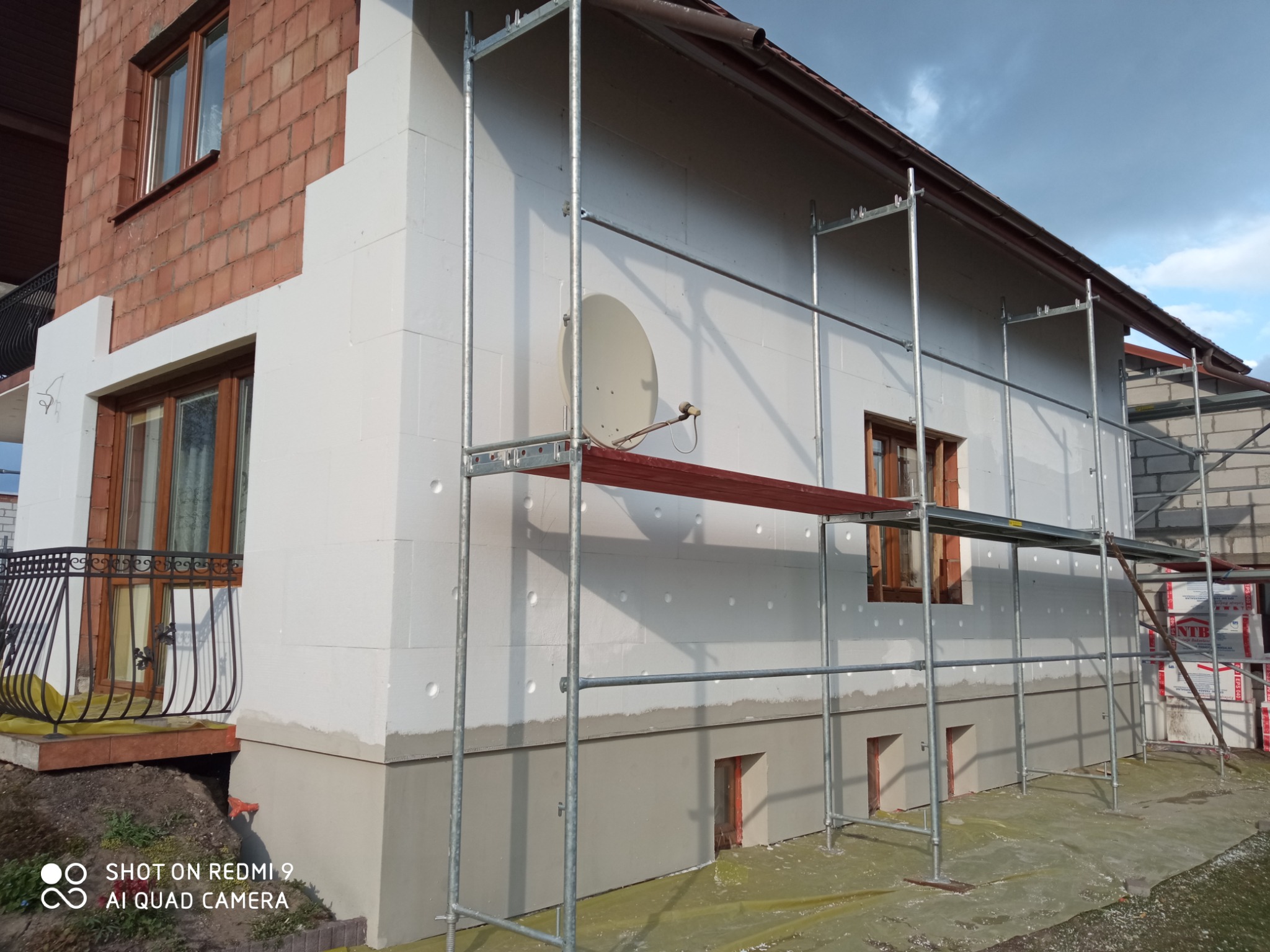 Dom w trakcie ocieplania styropianem, widoczne rusztowania, fragment elewacji wykończony czerwoną cegłą, antena satelitarna na ścianie.