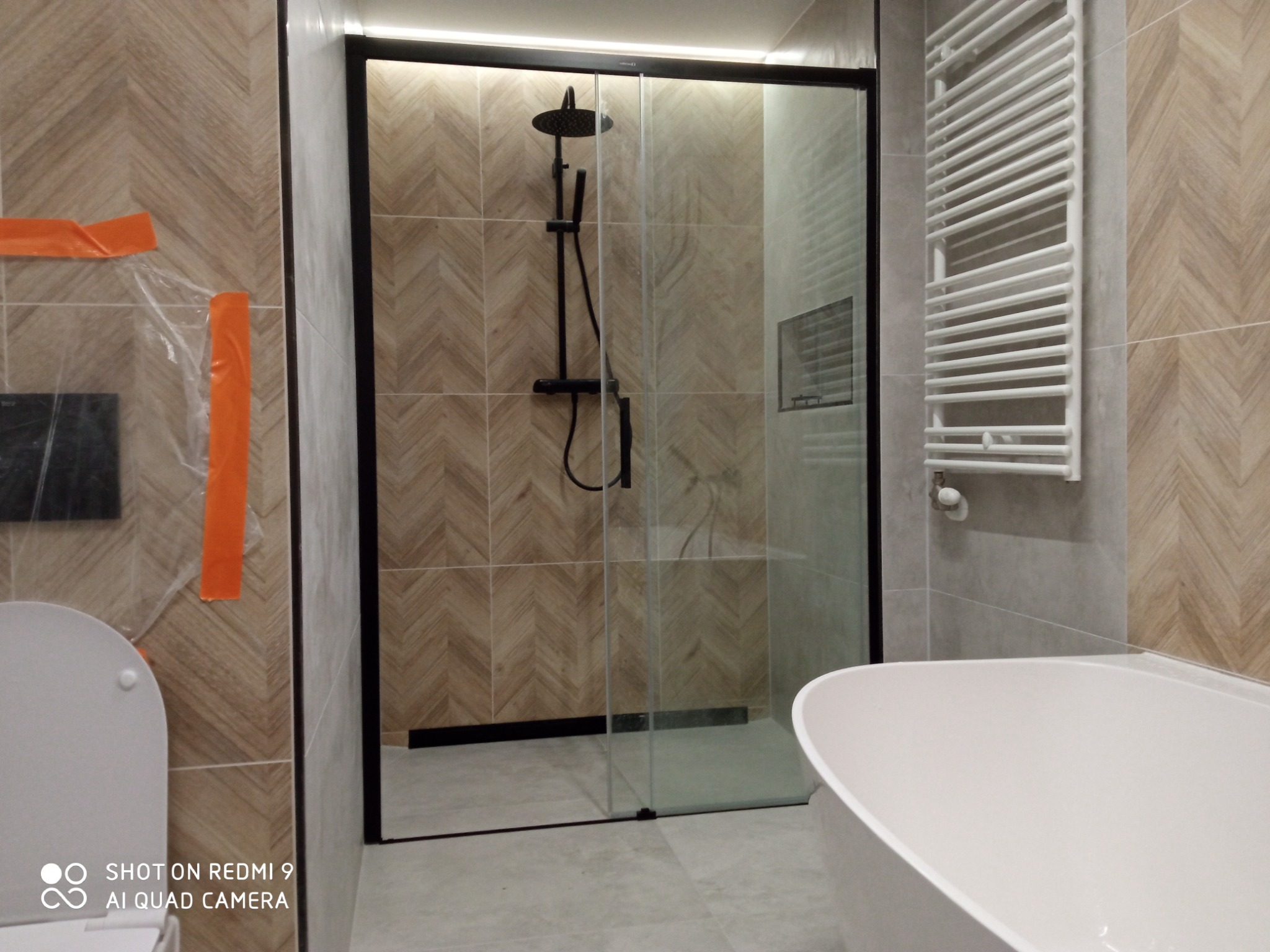 Nowoczesna łazienka z kabiną prysznicową typu walk-in z czarnymi elementami, grzejnikiem drabinkowym i częściowo widoczną wanną wolnostojącą, wykończona płytkami w geometryczny wzór jodełki,...