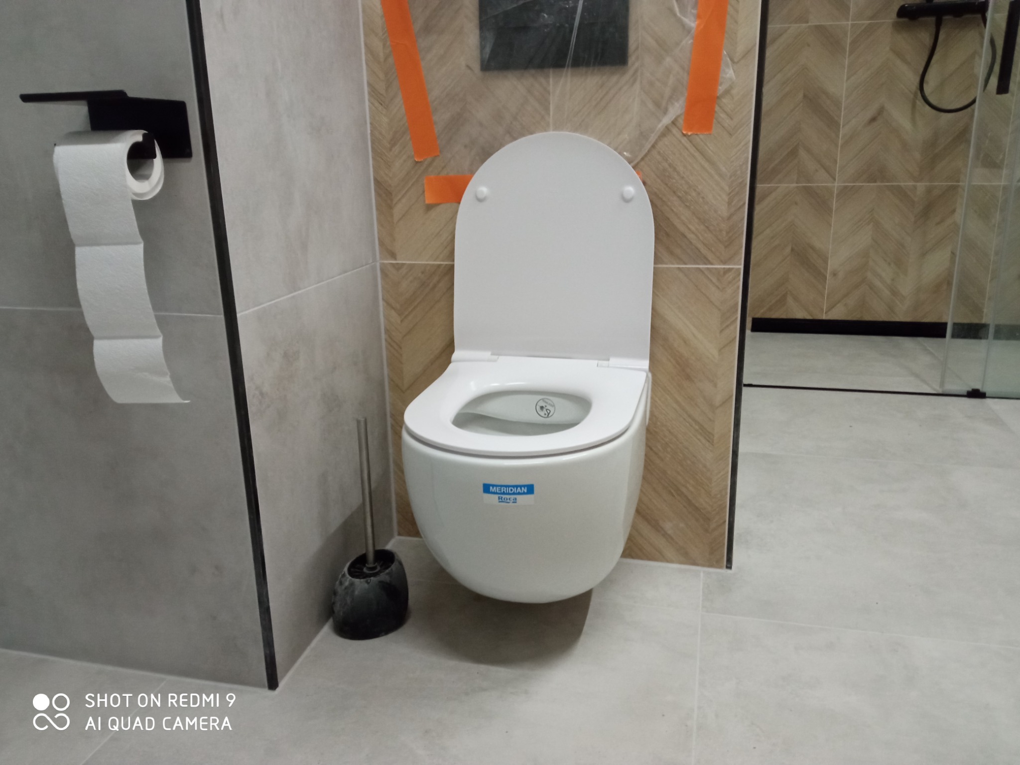 Nowoczesna toaleta podwieszana z deską sedesową, szczotką do WC i uchwytem na papier toaletowy w wykończonej łazience z płytkami w odcieniach szarości i beżu, zabezpieczona folią malarską.