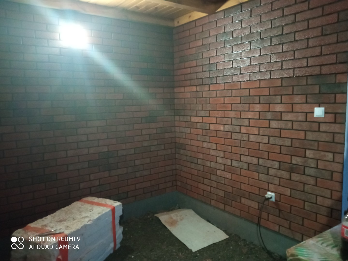 Wnętrze pomieszczenia z wykończoną ścianą imitującą czerwoną cegłę, widoczny fragment sufitu, biały włącznik światła, gniazdko elektryczne i materiały budowlane na podłodze.