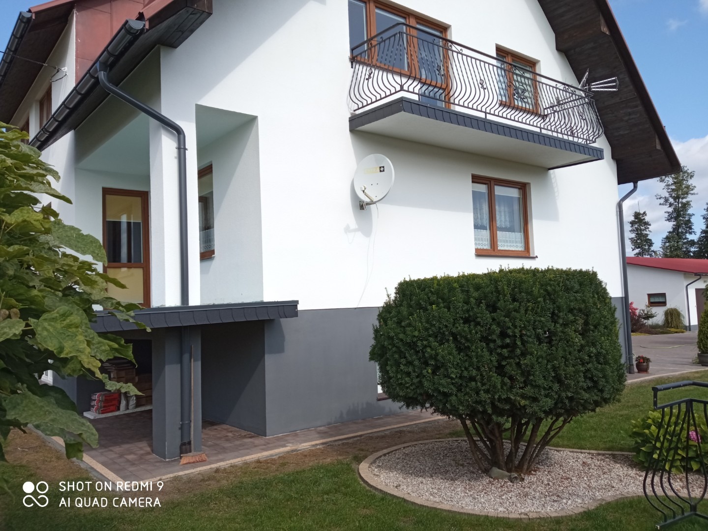 Dwupiętrowy dom z białymi ścianami, brązowymi oknami i balkonem z czarną balustradą, widok z zewnątrz na elewację i starannie przycięty krzew przed domem.