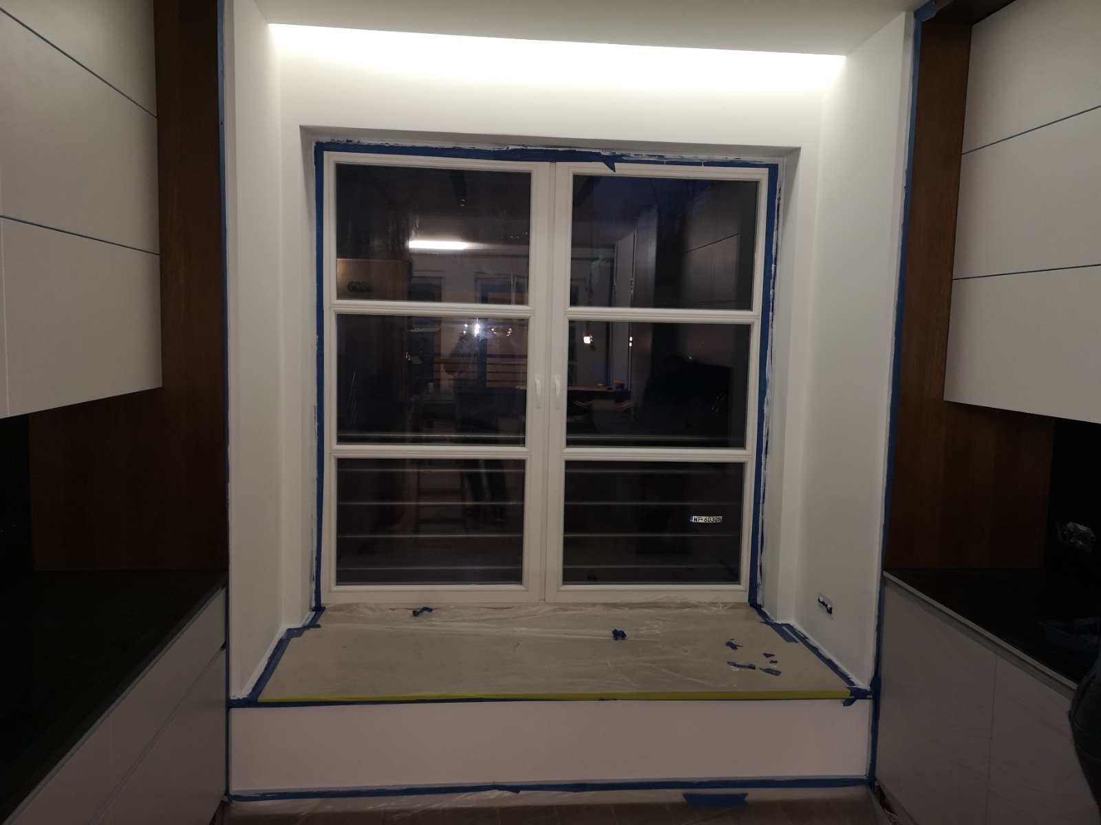 Przygotowanie do malowania wnęki okiennej z białym oknem, zabezpieczonej niebieską taśmą malarską i folią ochronną, widoczne fragmenty zabudowy kuchennej.