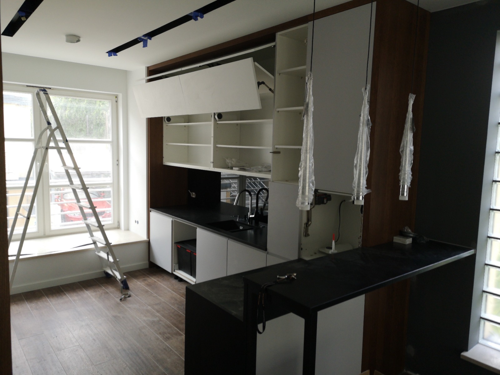 Wnętrze kuchni w trakcie montażu: białe szafki wiszące i stojące z czarnym blatem, surowe lampy wiszące w folii ochronnej, drabina oparta o okno, drewniana podłoga.