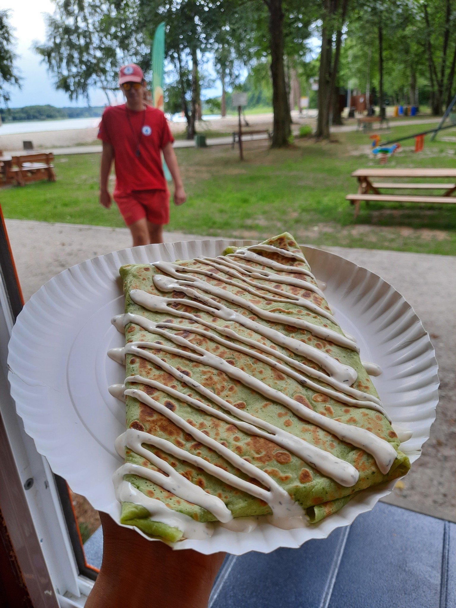 Zielony naleśnik z sosem na papierowym talerzu, w tle ratownik na plaży.