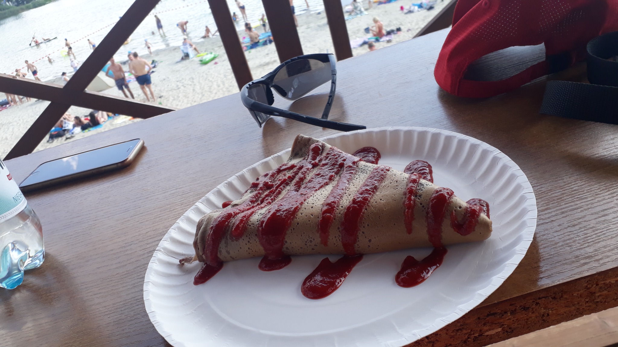 Naleśnik polany sosem truskawkowym na papierowym talerzu, na drewnianym stole z widokiem na plażę i jezioro w tle, obok leżą okulary przeciwsłoneczne, telefon i woda mineralna.