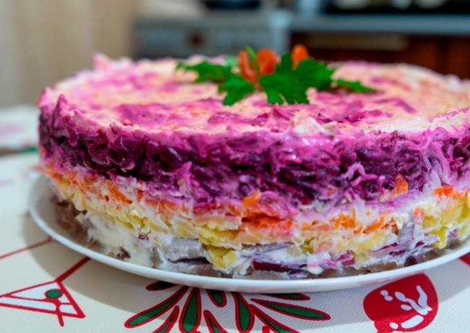 Warstwowa sałatka śledziowa udekorowana natką pietruszki i marchewką, podana na białym talerzu, na stole z wzorzystym obrusem.