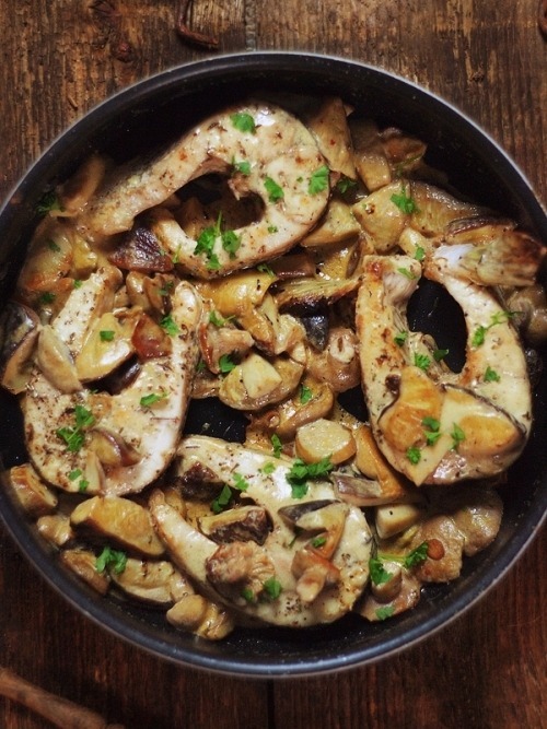 Smażone steki z ryby z grzybami w sosie śmietanowym, posypane świeżą pietruszką, w czarnej patelni na rustykalnym drewnianym stole.