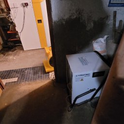 Osuszanie to My. - Urządzenia Trotec TTK 170 S i wentylator w piwnicy z wilgotną ścianą. Sprzęt do usuwania wilgoci w trakcie pracy. Widoczne przewody i elementy instalacji.