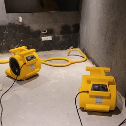 Osuszanie to My. - Dwa żółte wentylatory przemysłowe na szarym betonie, z widocznymi śladami wilgoci na ścianie i gniazdkiem elektrycznym. Sprzęt do usuwania wilgoci w pomieszczeniu.