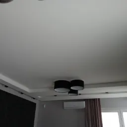 Nowoczesny sufit podwieszany z oświetleniem punktowym i centralną lampą z czarnymi abażurami, klimatyzacja na ścianie, ciemna ściana akcentowa z marmurkowym wzorem.