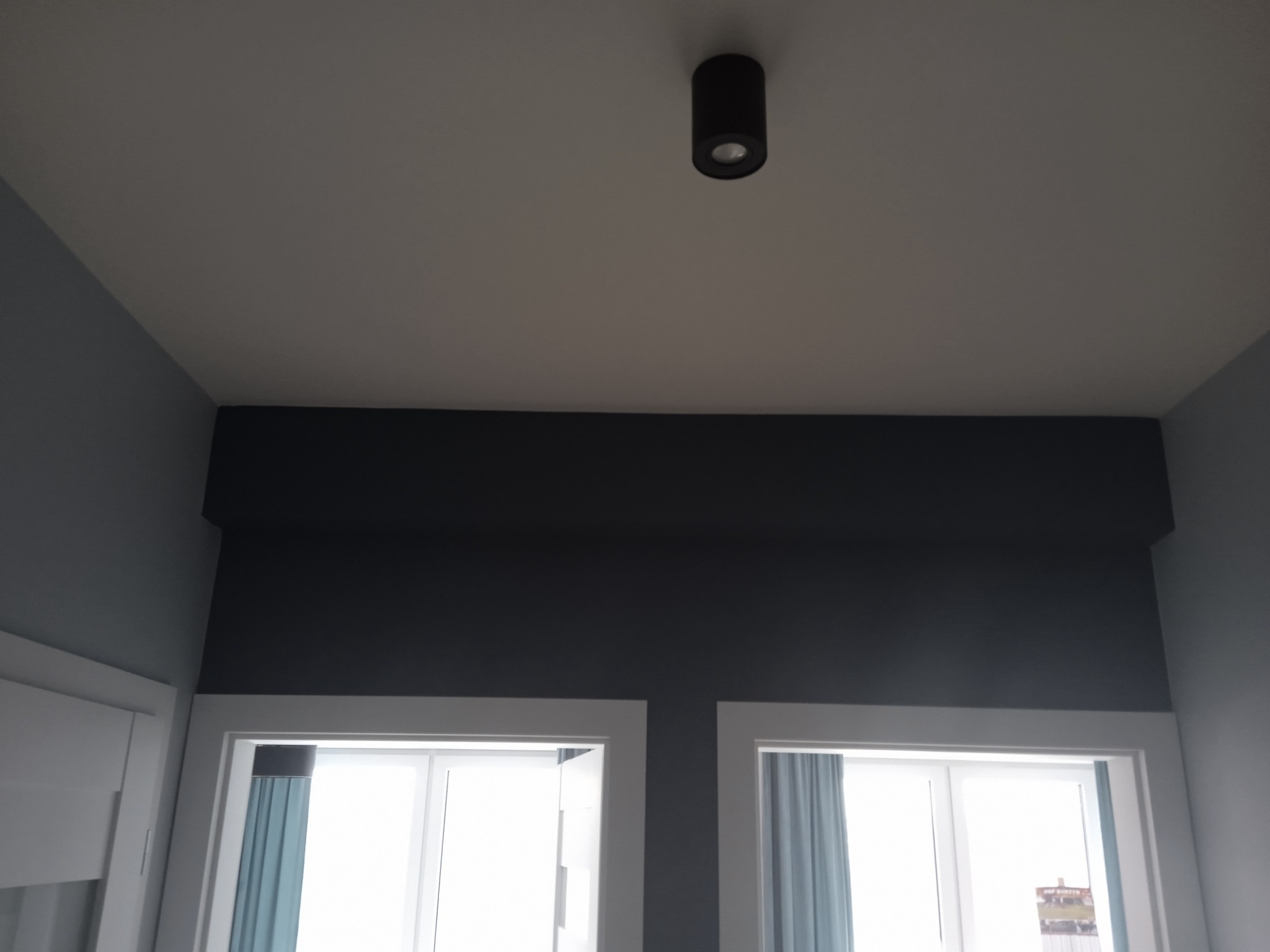 Fragment wnętrza z pomalowanymi na szaro ścianami nad dwoma oknami z białymi ramami i zasłonami, widoczna również czarna lampa sufitowa.
