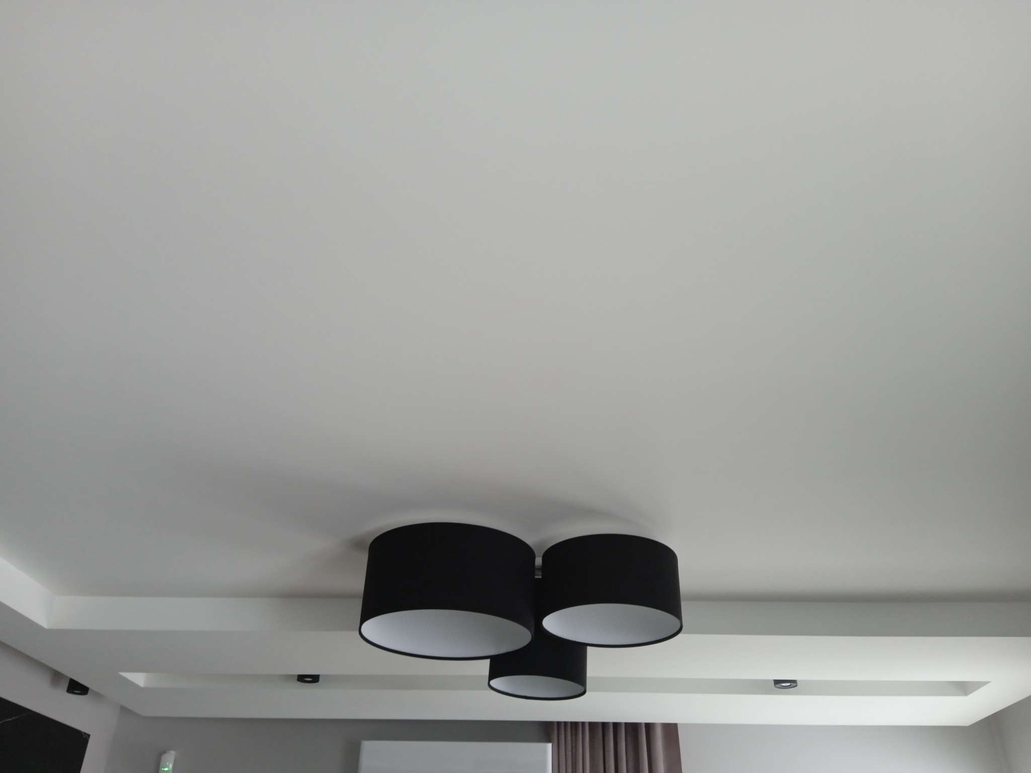 Trzy czarne, cylindryczne lampy sufitowe z białym wnętrzem kloszy, zamontowane na białym suficie podwieszanym z wbudowanym oświetleniem punktowym.
