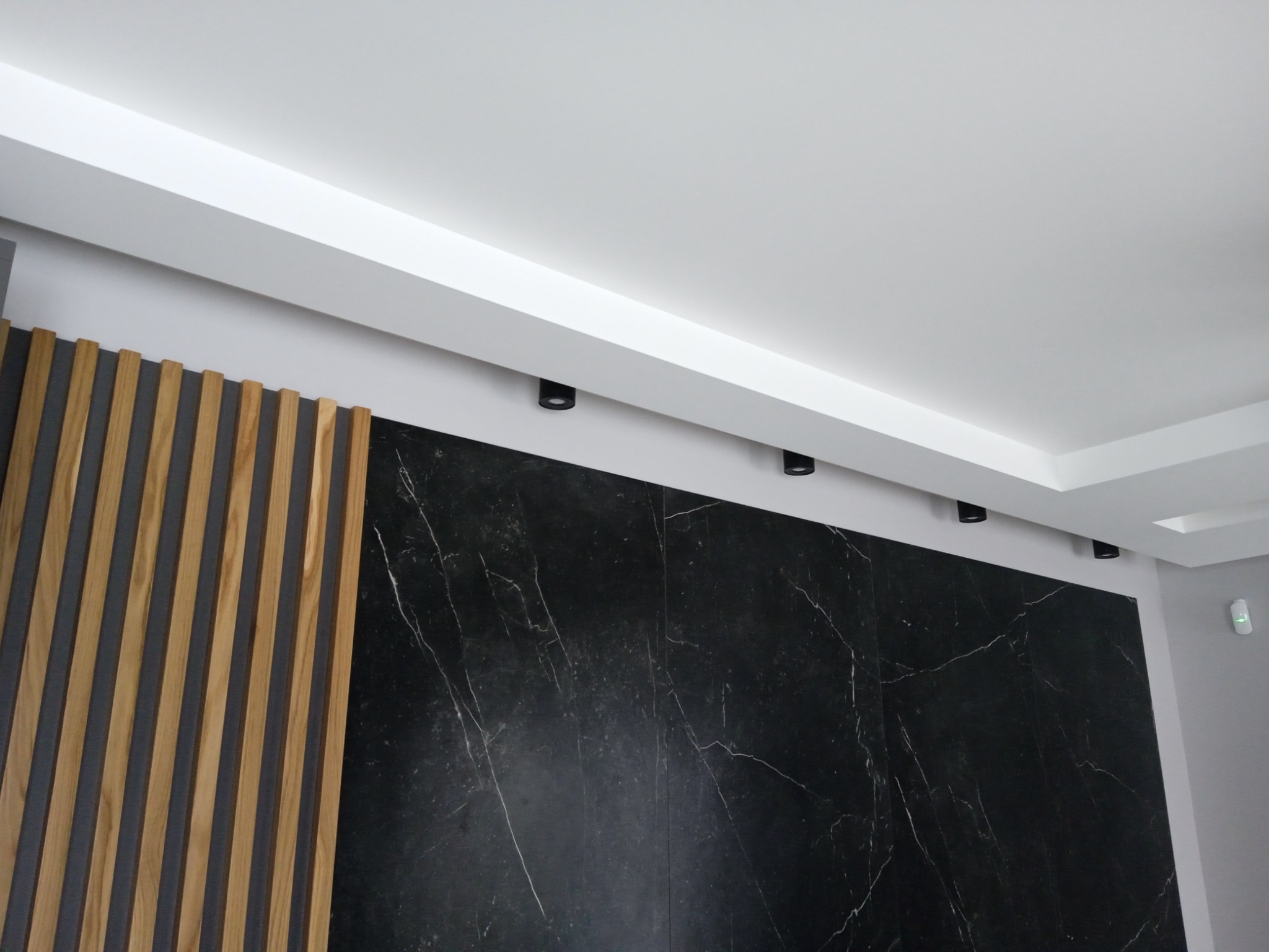 Wykończona ściana z czarnego marmuru z białymi żyłami, drewniane lamele pionowe po lewej stronie, sufit z oświetleniem punktowym.