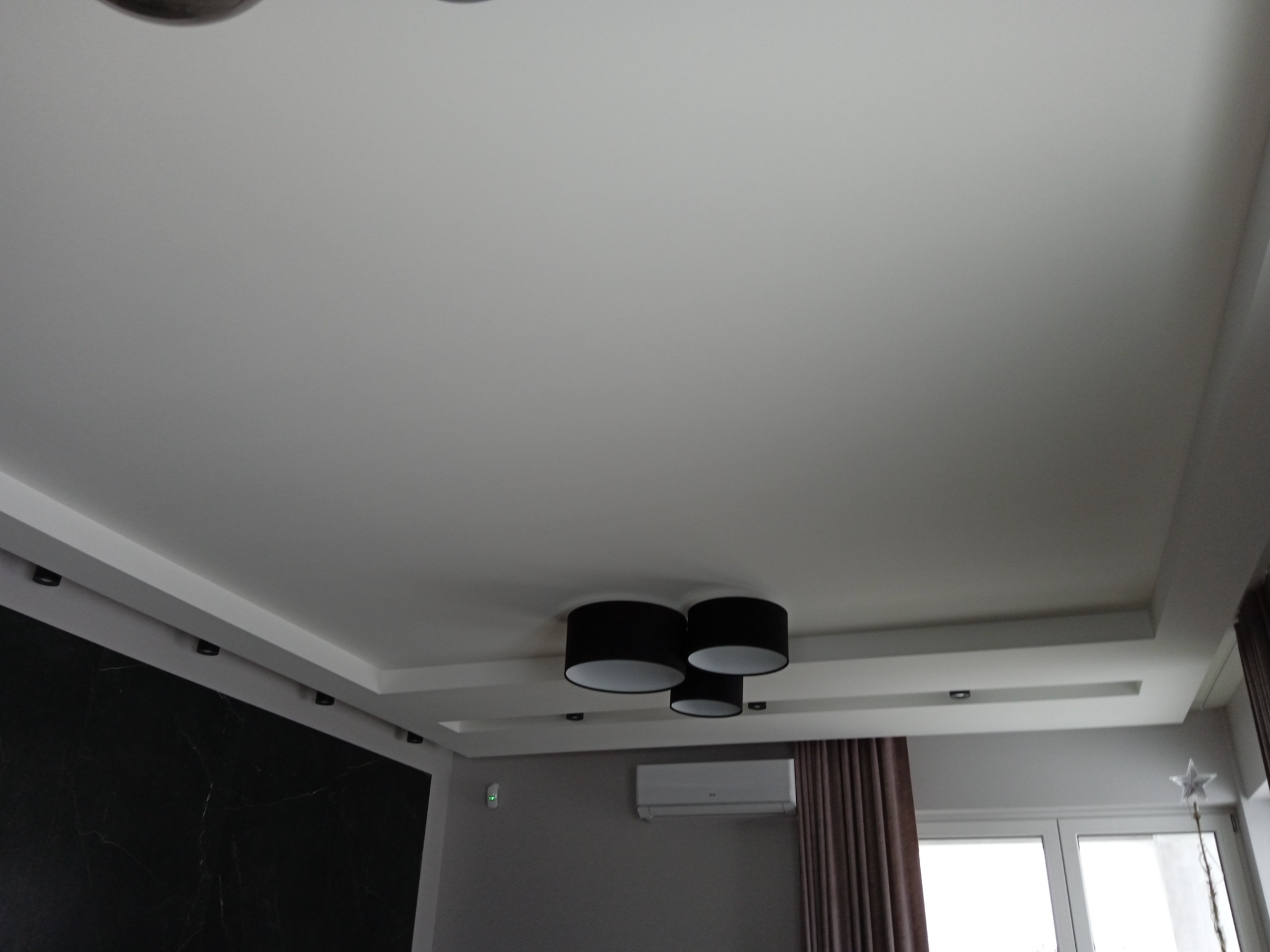 Nowoczesny sufit podwieszany z oświetleniem punktowym i centralną lampą z czarnymi abażurami, klimatyzacja na ścianie, ciemna ściana akcentowa z marmurkowym wzorem.