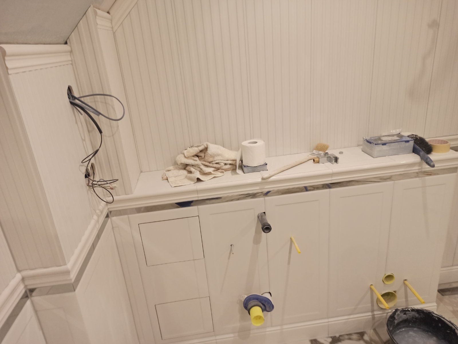 Biała łazienka w trakcie prac wykończeniowych: widoczne przewody elektryczne, otwory na rury, narzędzia i materiały budowlane na półce, pionowe panele ścienne.