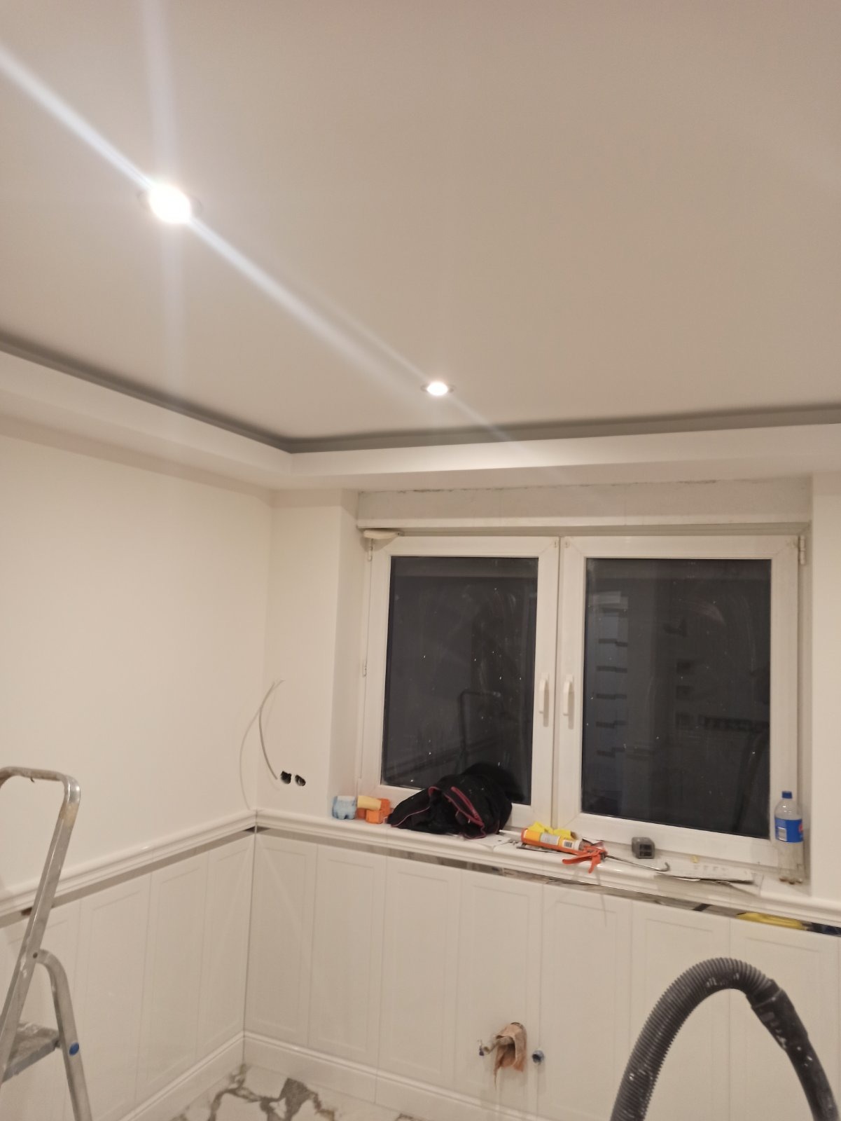 Wnętrze w trakcie prac wykończeniowych: białe ściany z dekoracyjną listwą, marmurowa podłoga, okno z narzędziami na parapecie, drabina oparta o ścianę, widoczne punkty oświetleniowe w suficie.