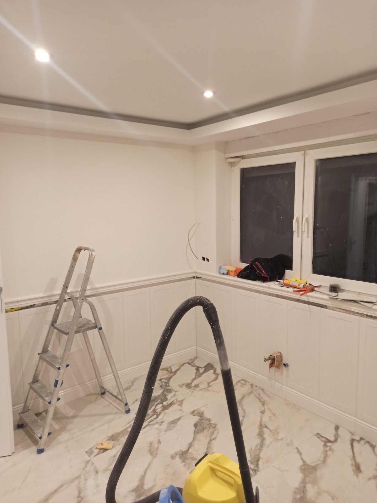 Pokój w trakcie prac wykończeniowych: białe ściany z panelami, marmurowa podłoga, drabina aluminiowa, odkurzacz przemysłowy i narzędzia na parapecie okna.