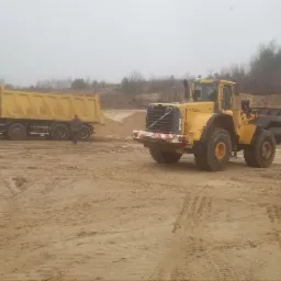 Żółta ładowarka Volvo L150F i dwie ciężarówki wywrotki na placu budowy z hałdami piasku w tle, mglisty dzień.