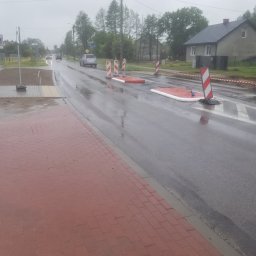 GPROJEKT Sp. z o.o. - Mokra asfaltowa jezdnia z pomarańczowo-białymi barierami i pachołkami, obok świeżo ułożony chodnik z czerwonej ceglanej nawierzchni. W tle ruch uliczny i zabudowania.
