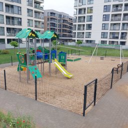 GPROJEKT Sp. z o.o. - Nowoczesny plac zabaw z kolorowymi zjeżdżalniami i huśtawkami na piaszczystym podłożu, otoczony siatkowym ogrodzeniem, obok chodnik z kostki. W tle białe bloki mieszkalne.