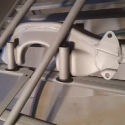 Elementy metalowe zawieszenia samochodowego po obróbce, pomalowane proszkowo na srebrno, umieszczone na metalowym stojaku.