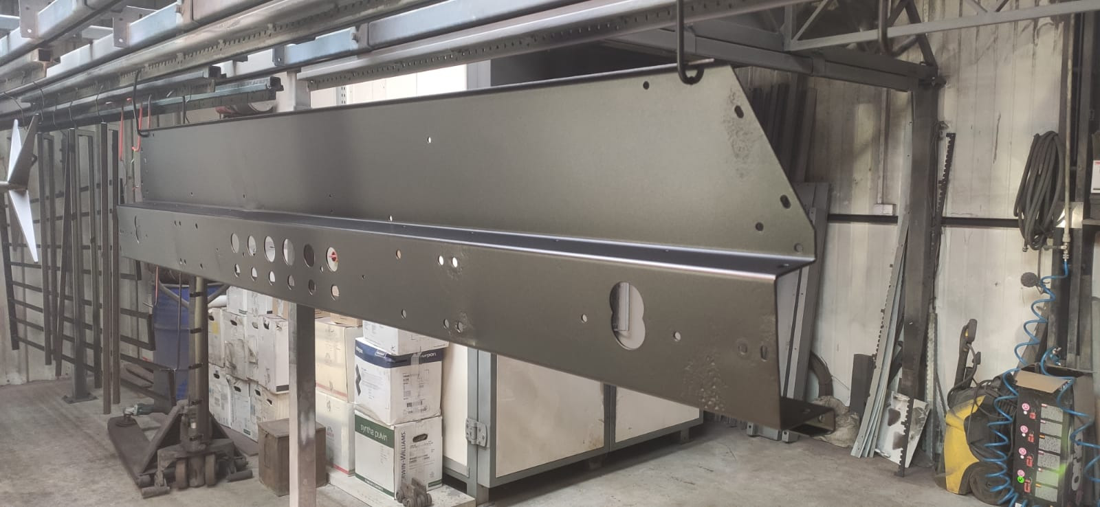Metalowy element konstrukcyjny z otworami, zawieszony na szynie w hali produkcyjnej, po procesie malowania proszkowego na kolor szary, z widocznymi zaciekami farby w dolnej części.
