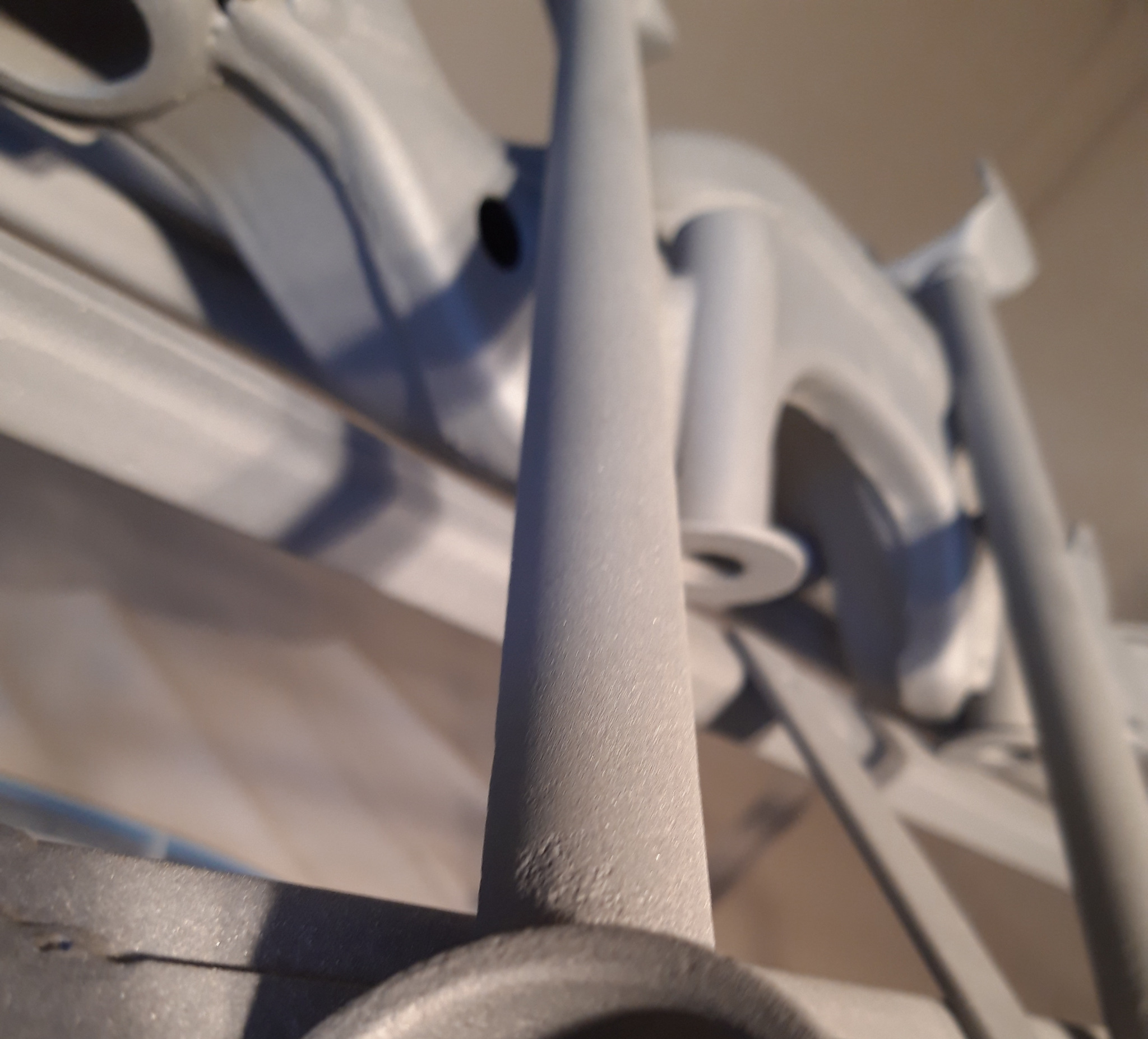 Metalowa konstrukcja po obróbce, pokryta jednolitą, jasnoszarą powłoką. Widoczne elementy rurowe i płaskie, z bliska, z efektem ziarnistości powierzchni. Nierównomierne oświetlenie podkreśla fakturę.