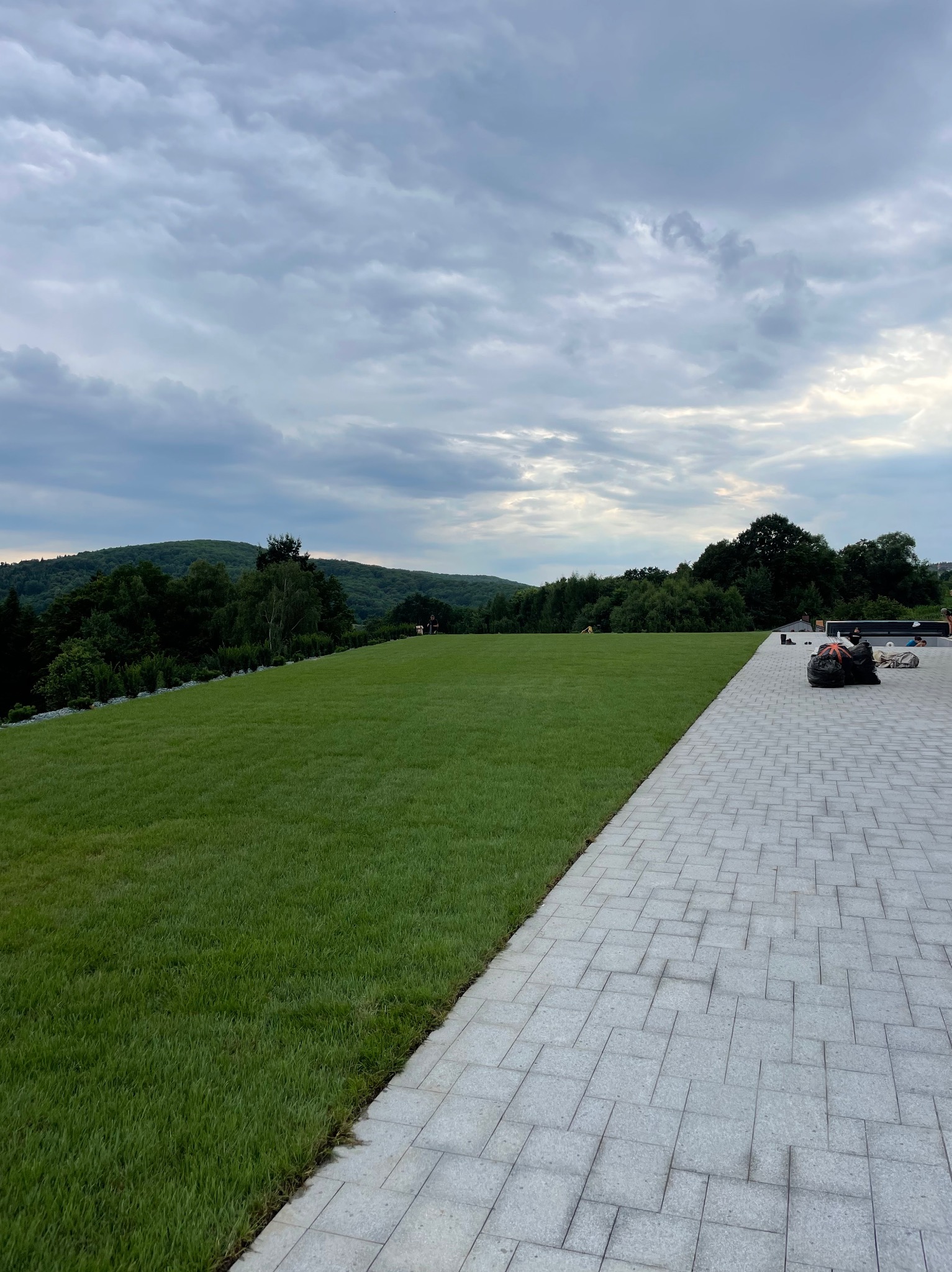 Świeżo ułożony trawnik z rolki sąsiadujący z szarą kostką brukową, w tle zalesione wzgórza pod pochmurnym niebem.
