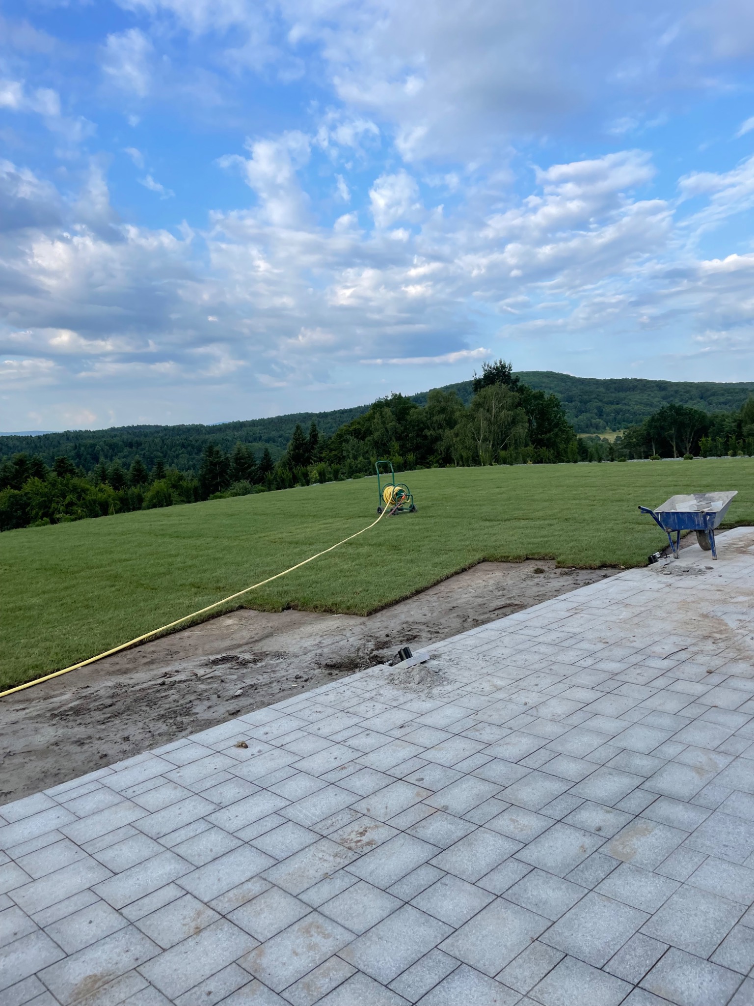 Układanie trawy z rolki na zboczu, widok na zielone wzgórza, w tle las, wąż ogrodowy i taczka na trawniku, częściowo ułożona kostka brukowa na pierwszym planie.