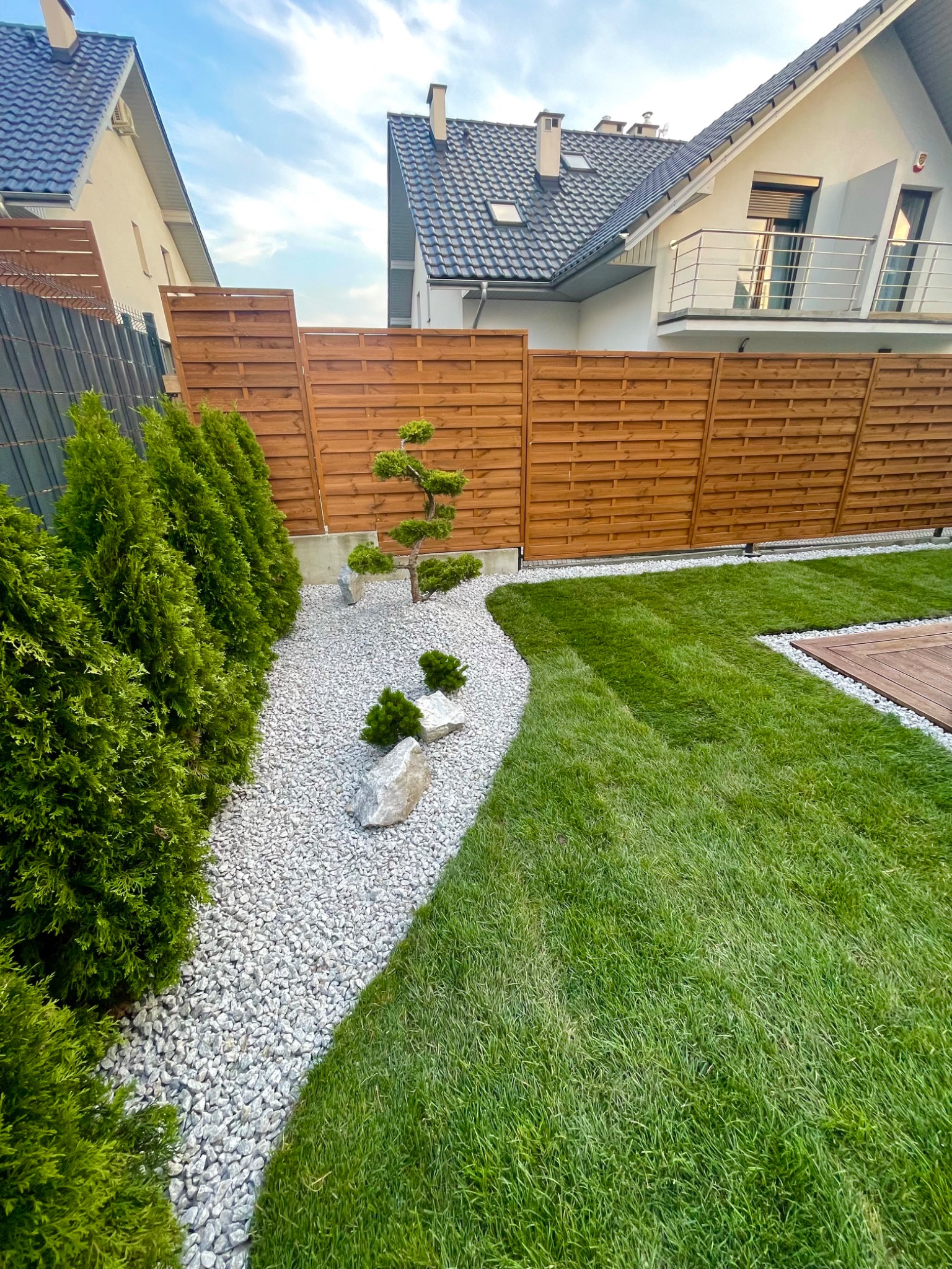 Starannie zaprojektowany ogród z równo przystrzyżonym trawnikiem, ścieżką z białego żwiru, ozdobnymi krzewami i formowanym drzewkiem bonsai na tle drewnianego ogrodzenia.