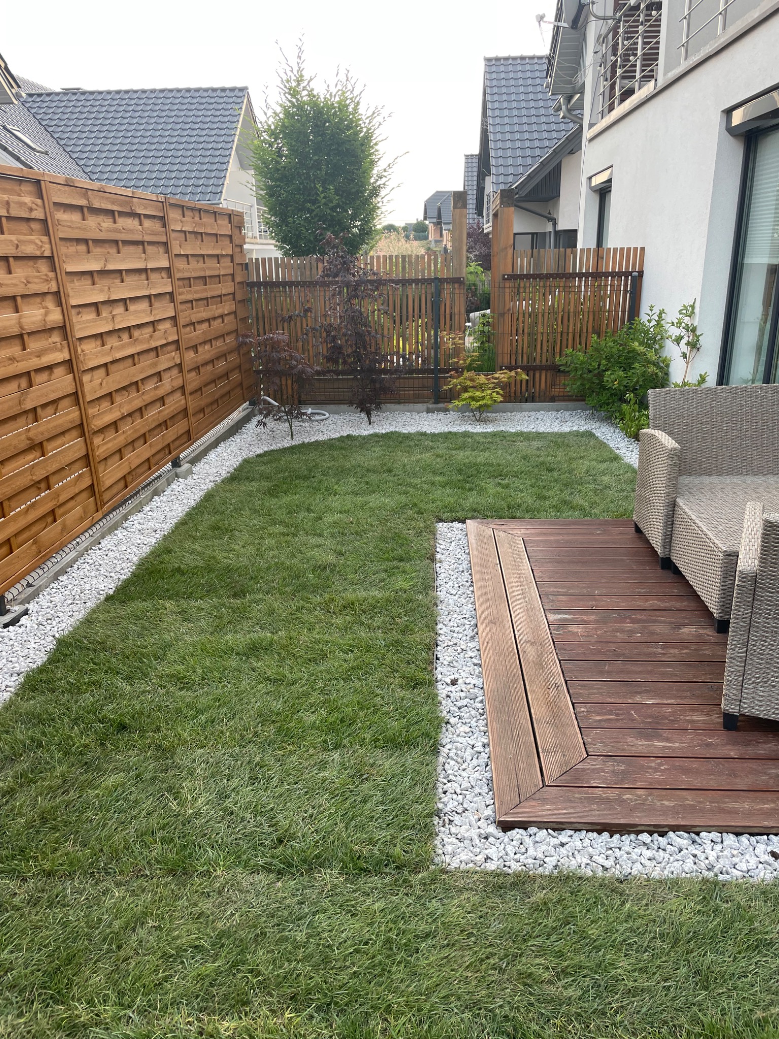 Starannie zagospodarowany przydomowy ogród z równo przystrzyżoną trawą, drewnianym tarasem, meblami ogrodowymi z rattanu, ozdobnymi drzewkami i białymi kamykami oddzielającymi trawnik od ogrodzenia.