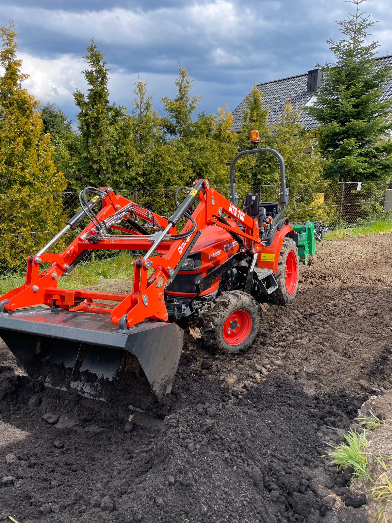 Pomarańczowy traktor Kubota MTS 700 z łychą spulchnia ziemię na polu pod uprawę, w tle zielone drzewa i dom pod pochmurnym niebem.