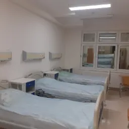 Wnętrze nowocześnie urządzonej sali szpitalnej z trzema łóżkami, szafkami nocnymi i klimatyzacją, widok przez okno na inną salę.